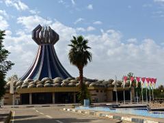 A unique building with a cone-shaped tower and boulder-like facade stands under a blue sky with wispy clouds, next to a palm tree and a row of flags.
