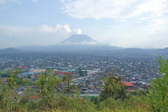 Circuit 1: Gorillas-Chimps-Nyiragongo-Pygmies-Okapi-Kigali – 7 Days A wide shot of a densely populated city sprawling out towards a large mountain, partly obscured by clouds, under a bright sky.
