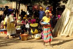 A vibrant gathering of people, predominantly women, dressed in colorful fabrics and headwraps, fills an outdoor space near a building and a large tree.