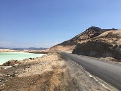 A road curves alongside a light green lake, leading towards a rocky mountain under a clear blue sky.
