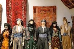 A display of mannequins dressed in traditional Kurdish clothing, standing in front of vibrantly colored rugs.