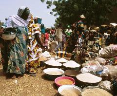 A bustling African marketplace shows women in colorful clothing selling grains from bowls and bags amidst a crowd.