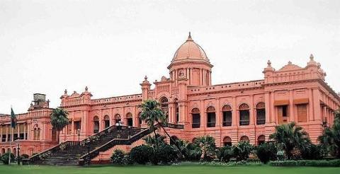 This is a frontal view of the Ahsan Manzil, a pink palace with a grand dome and staircase, surrounded by green lawns and trees, under a cloudy sky.