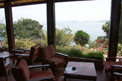 Eine Veranda mit gestreiften Stühlen und Tischen bietet Aussicht auf üppiges Grün und ein Gewässer in der Ferne und schafft so eine ruhige und einladende Atmosphäre.