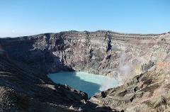 A panoramic shot shows a volcanic crater filled with turquoise water, surrounded by layered rock formations under a clear blue sky.