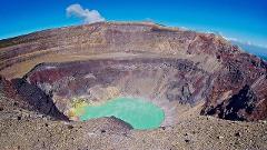 A stunning view of a massive volcanic crater with a turquoise lake at its bottom, surrounded by colorful rock formations under a bright blue sky.