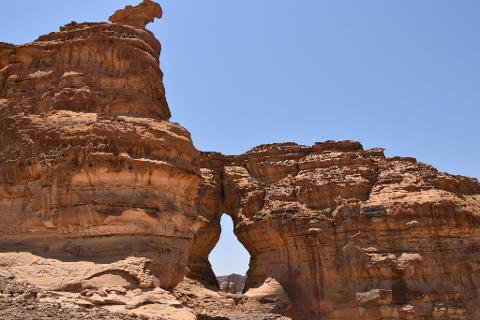 This is a landscape of weathered, reddish-brown rock formations with a natural archway carved through the stone, against a clear blue sky.