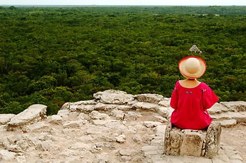 A woman in a red dress and hat sits on ancient stone ruins overlooking a vast, dense green jungle.