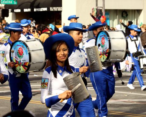 A vibrant parade with musicians in blue and white uniforms, playing drums and guiros, the woman in the front smiling confidently.