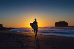 A silhouetted surfer walks along a sandy beach at sunset, carrying a surfboard, with waves crashing and a rock formation visible in the background.