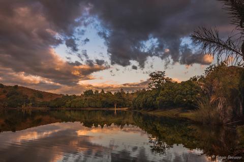 A serene landscape featuring a lake reflecting a colorful sky with dramatic clouds, lush greenery, and trees along the shoreline, creating a tranquil atmosphere.