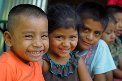 A group of smiling, young children of Latin American descent stand closely together, looking directly at the camera.