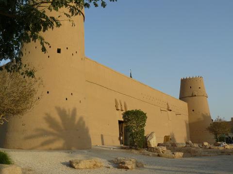 A sunny, low-angle shot showcases Al Masmak Fort in Riyadh, highlighting its tall, cylindrical towers and the long, thick wall, with plants and rocks in the foreground.