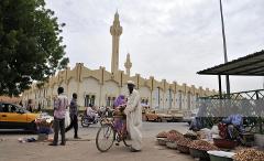 A street scene in N'Djamena, Chad, with a mosque in the background, people walking, a bicycle vendor, and market stalls selling nuts.