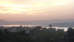 A serene sunset bathes a landscape with a wide river winding through lush trees, with distant hills and soft, peach-colored sky adding to the peaceful atmosphere.