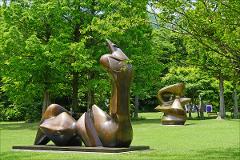 Two bronze abstract sculptures sit on a grassy lawn surrounded by lush green trees, creating a serene outdoor art display.
