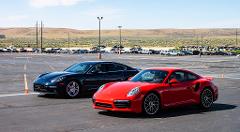 A black Porsche Panamera and a red Porsche 911 Turbo parked in a parking lot dotted with orange cones, with other cars and a desert landscape in the background.
