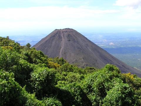 A volcano with a trail visible up its side, surrounded by green vegetation, under a clear blue sky.