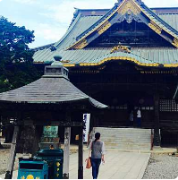 En kvinde går op ad trappen mod en stor, udsmykket japansk tempelbygning med et indviklet, lagdelt tag og gulddetaljer, mens en mindre pagodelignende struktur står til siden.