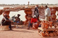 A group of people are gathered on a sandy area next to a body of water, with pottery items displayed on a table.