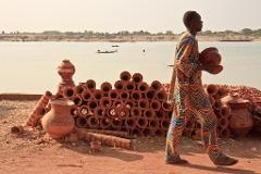 A man in colorful clothing carries a clay pot past stacks of pots by a river with boats.