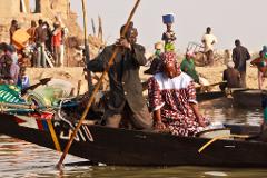 A man poles a boat through the water, transporting a woman in traditional dress and various goods, with a bustling shoreline of people in the background.