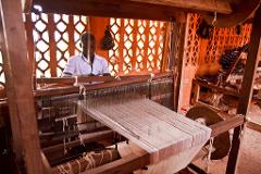 A man in a white shirt works at a wooden loom in a room with an orange wall and decorative windows.