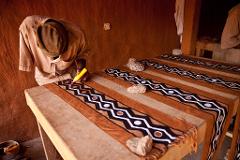 An artisan in a tan shirt and hat uses a yellow tool with red dye to paint black and white geometric patterns on a brown fabric, weighted down by stones.