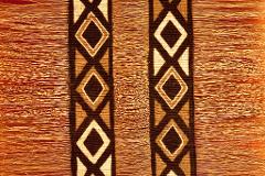 Close-up of a woven textile with diamond patterns in dark brown, beige, and cream, fringed with long, straw-colored strands.