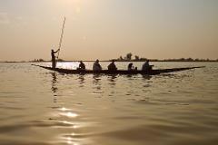 A long boat with a group of people is silhouetted on calm water, possibly at sunset.