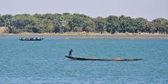 Two boats are sailing on a wide river; one boat is full of people, while the other is carrying one person and some sand.