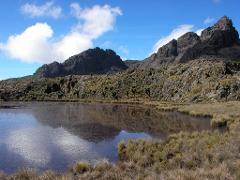A serene landscape with a reflective lake mirroring a rugged mountain range under a bright blue sky with scattered clouds.