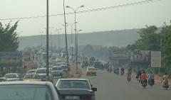 A busy street scene in an African city, with cars, motorcycles, and billboards lining the road, all under a hazy sky.