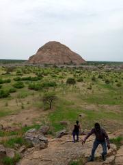A man and woman climb a rocky outcrop with a large, dome-shaped rock formation in the background, amidst a green, sparsely treed landscape under an overcast sky.