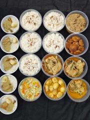 An overhead shot of 16 plastic containers filled with various food items on a black surface, including rice with fried onions, chicken and egg with rice, curried chicken, meatballs, and stewed vegetables and eggs.