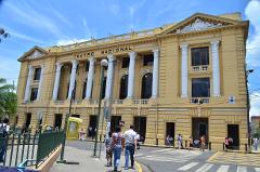 A large, yellow building with the words "Teatro Nacional" on top, tall white columns, and people walking on the sidewalk in front of it, under a blue sky with scattered clouds.