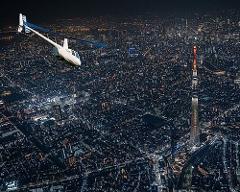 A helicopter flies over a densely populated city illuminated at night, with a tall tower standing out amidst the urban landscape.