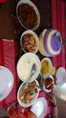 A table laden with various dishes, including rice, curries, and fried items, alongside plates and a thermos, set on red and white patterned mats.