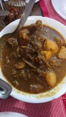 A close-up shot of a bowl of curry with meat and potatoes, along with a spoon and a small bowl of food in the background. It's placed on a red and white striped tablecloth.
