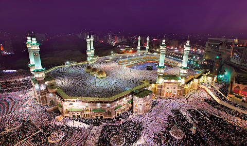 This is an aerial view of the Masjid al-Haram in Mecca at night, with a large crowd of people gathered for prayer around the Kaaba.