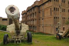 A weathered artillery gun stands prominently in the foreground, with a damaged building in the background, hinting at conflict or decay.