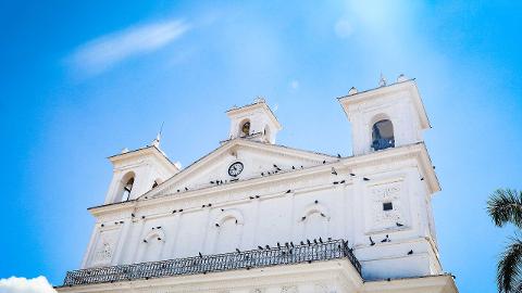 A white church with two bell towers stands tall against a bright blue sky, adorned with birds perched on its facade.