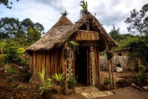 A traditional Papua New Guinean hut with a thatched roof and wooden walls stands amidst lush greenery, adorned with skeletal figures flanking its entrance.