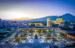 A scenic cityscape featuring a brightly lit central plaza with illuminated buildings and a mountain backdrop under a dusky sky.