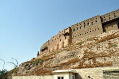 A grand brick structure stands atop a rugged hillside under a clear blue sky, overlooking a modest building and sparse vegetation at the base.