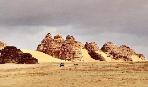 A car drives through a desert landscape with sandstone formations under a cloudy sky.