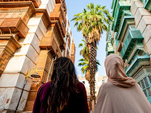 Two people stand between buildings with detailed wooden balconies in Jeddah's Al-Balad district, looking up at palm trees against a blue sky.