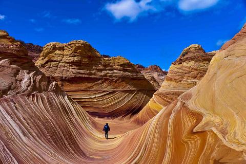 A lone figure walks through a canyon of layered, swirling sandstone formations under a bright blue sky.