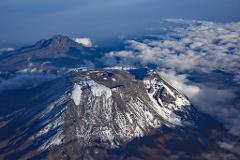 An aerial view captures the majestic snow-capped peak of Mount Kilimanjaro with a smaller mountain in the background and a sea of fluffy clouds below a clear blue sky.