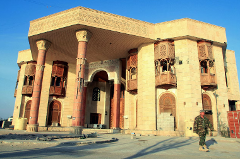 A grand, ornate building stands under a clear sky, featuring detailed columns, balconies, and arched windows, with a soldier in camouflage standing in front of it.